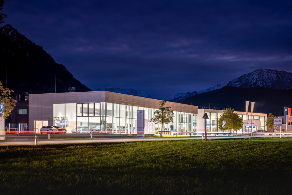 Referenz Holzbau Saurer Autohaus Schweiger Austellungshalle moderner Objektbau nachhaltige Architektur Verglasung Nachtaufnahme Beleuchtung eingebettet in Gebirge